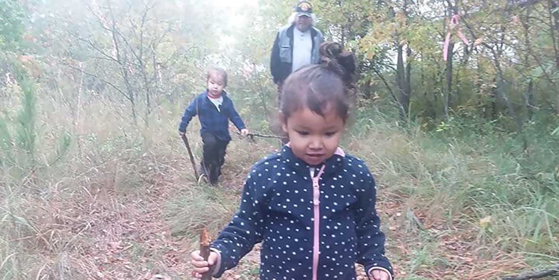 Two small Ojibwe children are joined by their grandfather on a walk through the woods.