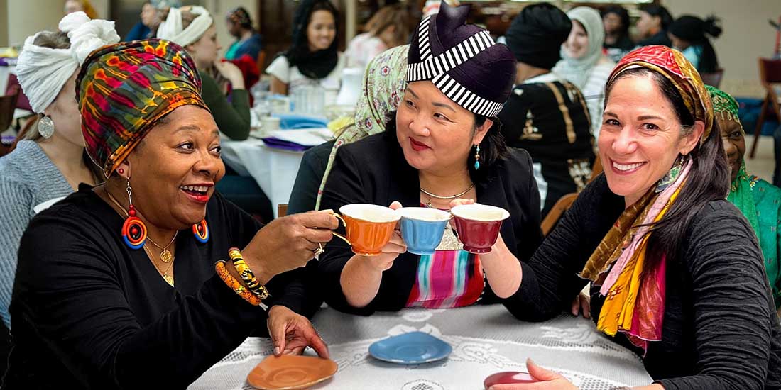 Three women in headscarves toast one another with brightly colored tea cups.